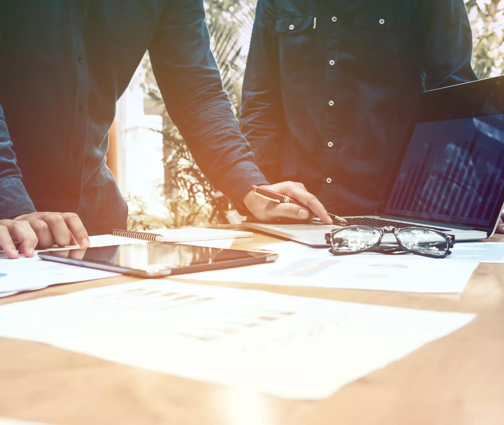 Three businessmen working on laptop computer with documents and paperwork scattered around and digital tablet on desk.