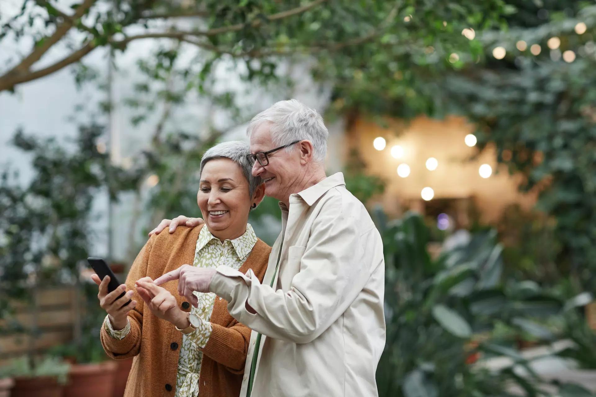 a couple standing in a garden area, smiling while looking together at a phone
