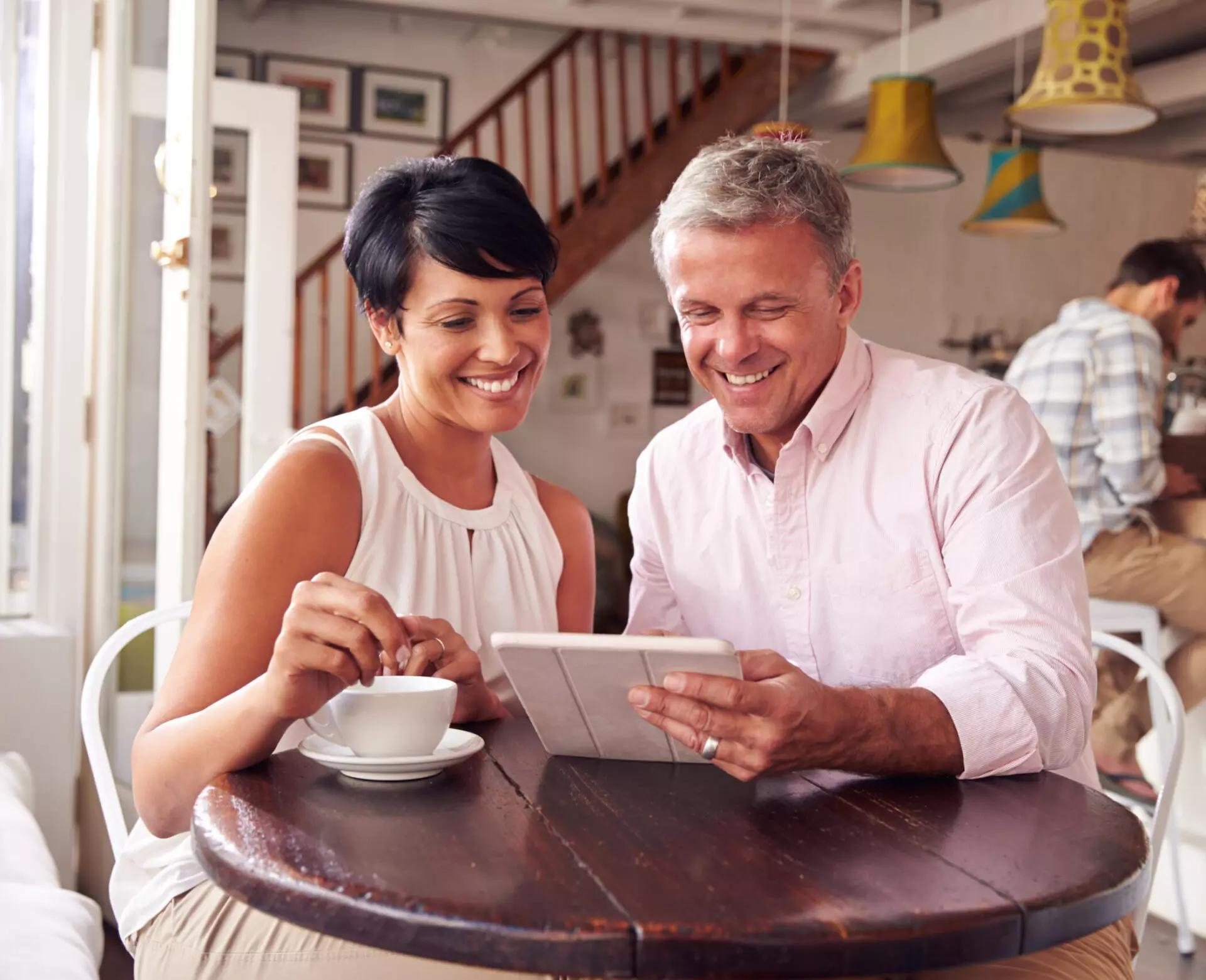 a man and a woman, both smiling, look at a tablet screen while sitting together at a table