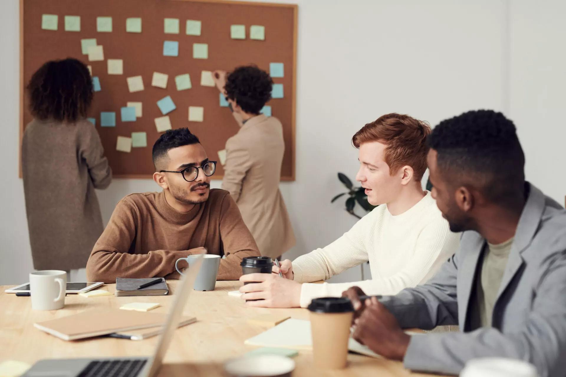 three people sitting at a table in an office while two other people adjust post it notes on a bulletin board in the background