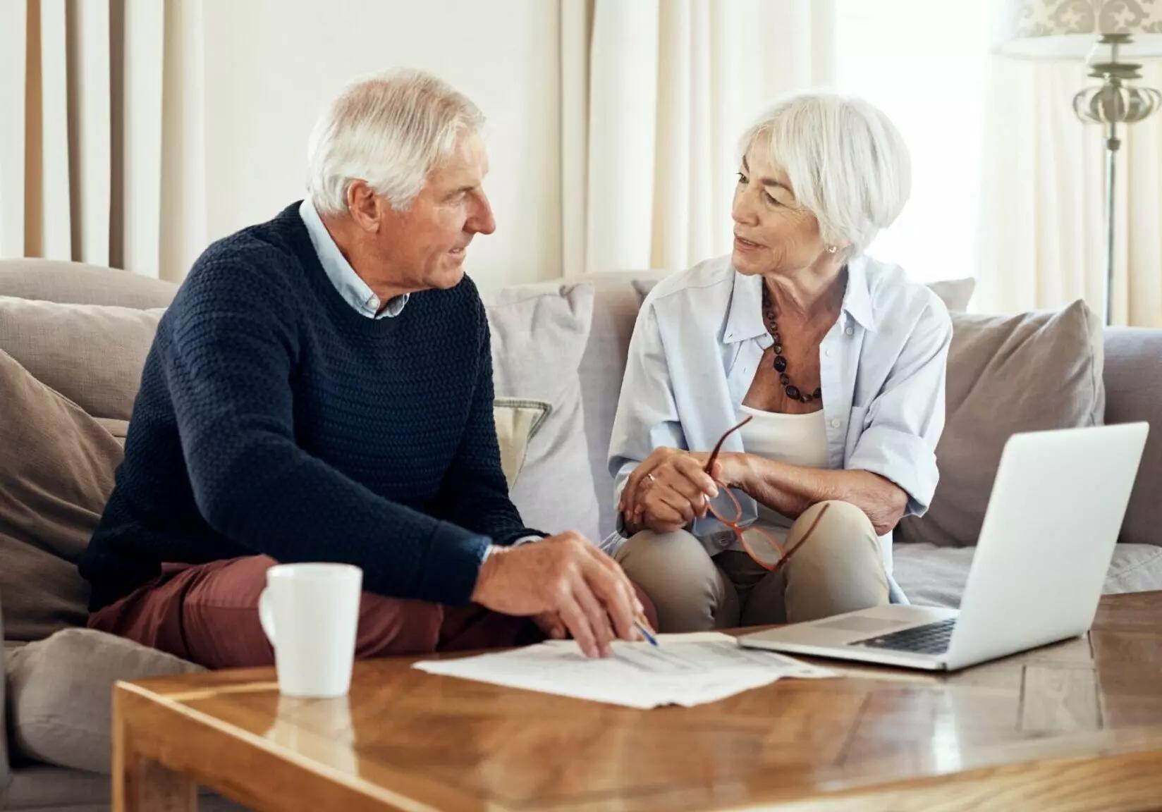 An older man and woman sit on a couch, in conversation, with papers and a laptop on a coffee table in front of them discussing fees
